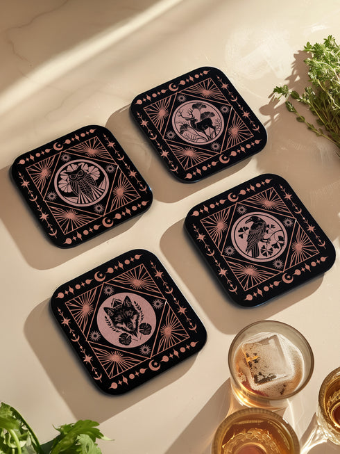 A set of four black, high-gloss square coasters featuring intricate rose-gold metallic illustrations of an owl, raven, stag, and wolf framed by celestial and botanical motifs. Made from durable masonite hardboard with cork backing for slip-resistant protection.